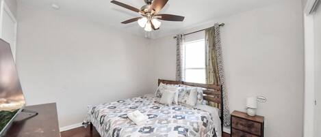 Un dormitorio con cama, ventilador de techo, ventana con cortinas, mesita de noche y escritorio.
