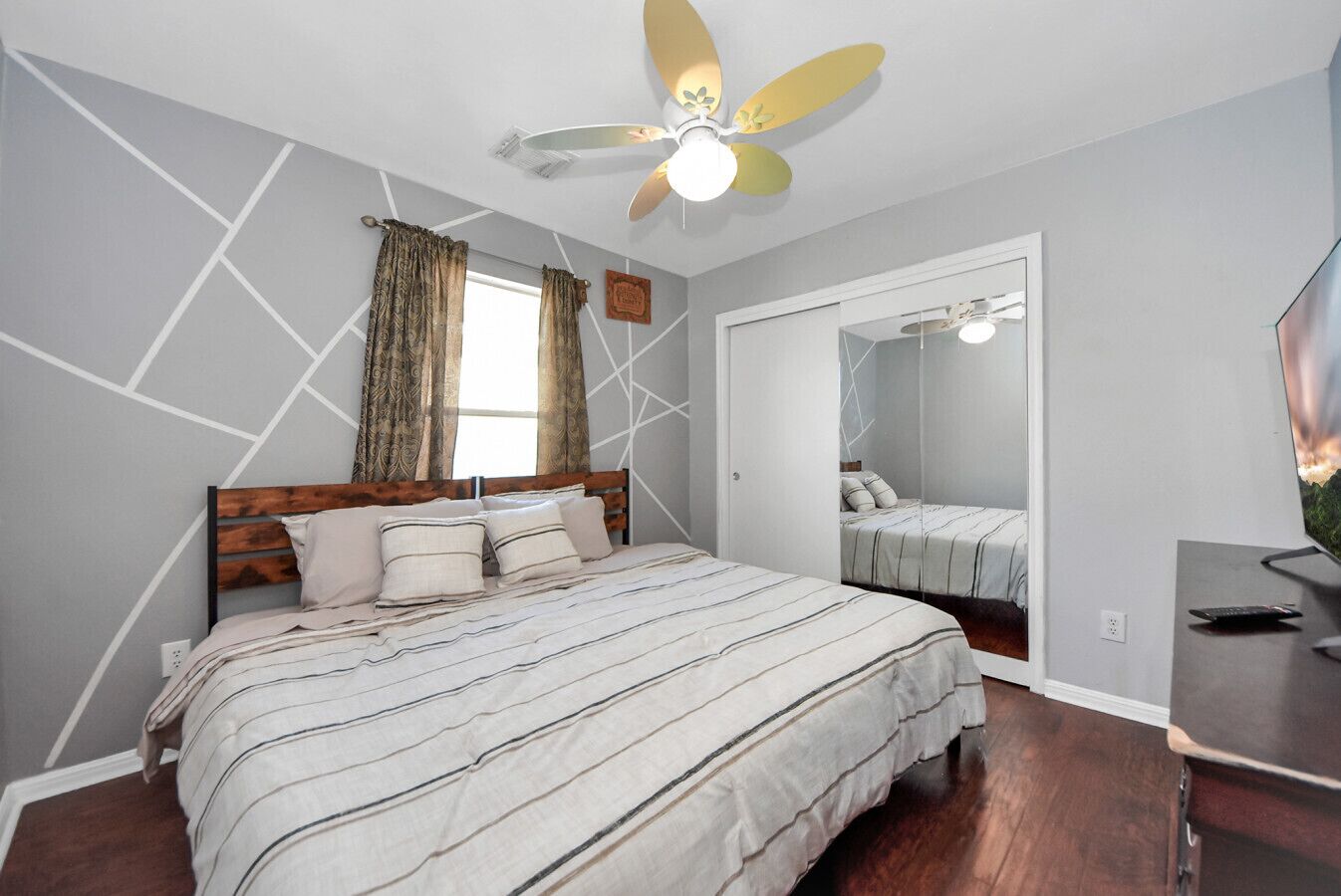Un dormitorio con cama, ventilador de techo, ventana con cortinas, espejo y televisor.