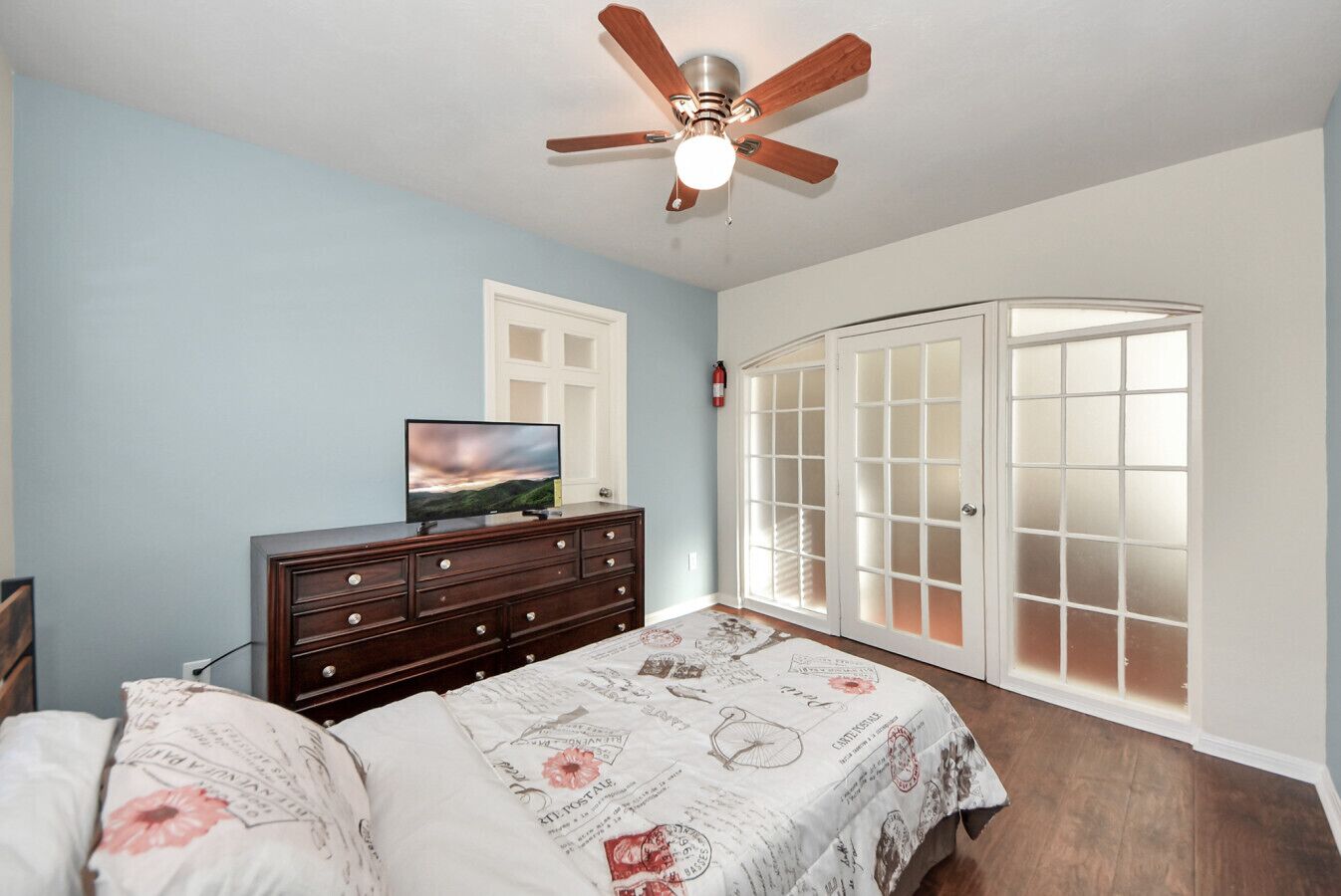 Une chambre à coucher comprenant un lit, une commode, un téléviseur, un ventilateur de plafond et une grande fenêtre dotée de portes en verre dépoli.