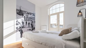 A bedroom with a large window, a bed with white bedding, a wall-mounted lamp, and a poster on the wall. - Noordzee Apartment 2 (Zandvoort)
