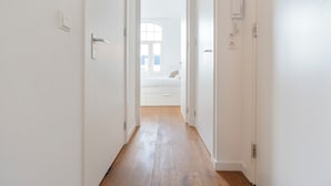 A hallway with white walls and wooden flooring leading to a bedroom with a bed and a window. - Noordzee Apartment 2 (Zandvoort)