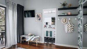 Una habitación con suelo de madera, una silla, una mesita, un espejo, un televisor, un armario y un baño visible a través de una puerta abierta.