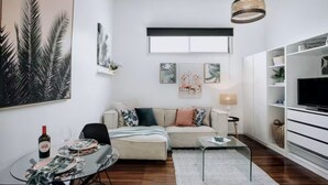 Una sala de estar con un sofá, una mesa de centro de vidrio, un televisor y un comedor con una mesa dispuesta para dos personas.