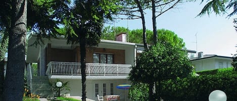 Una casa de dos pisos con un balcón, un jardín pequeño con una banca y una mesa con un paraguas colorido.
