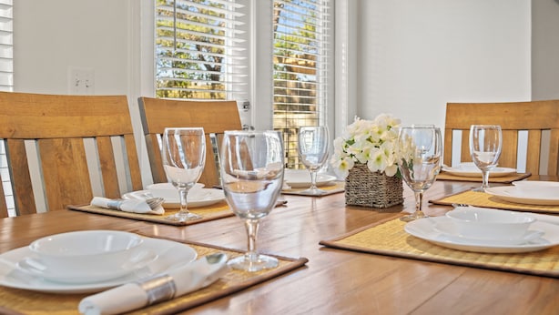 A dining table set with white plates, clear glasses, and a woven basket with white flowers. - The Winking Turtle – Your Shell-tered Beach Escape! Sleeps 10! (Inlet Beach)