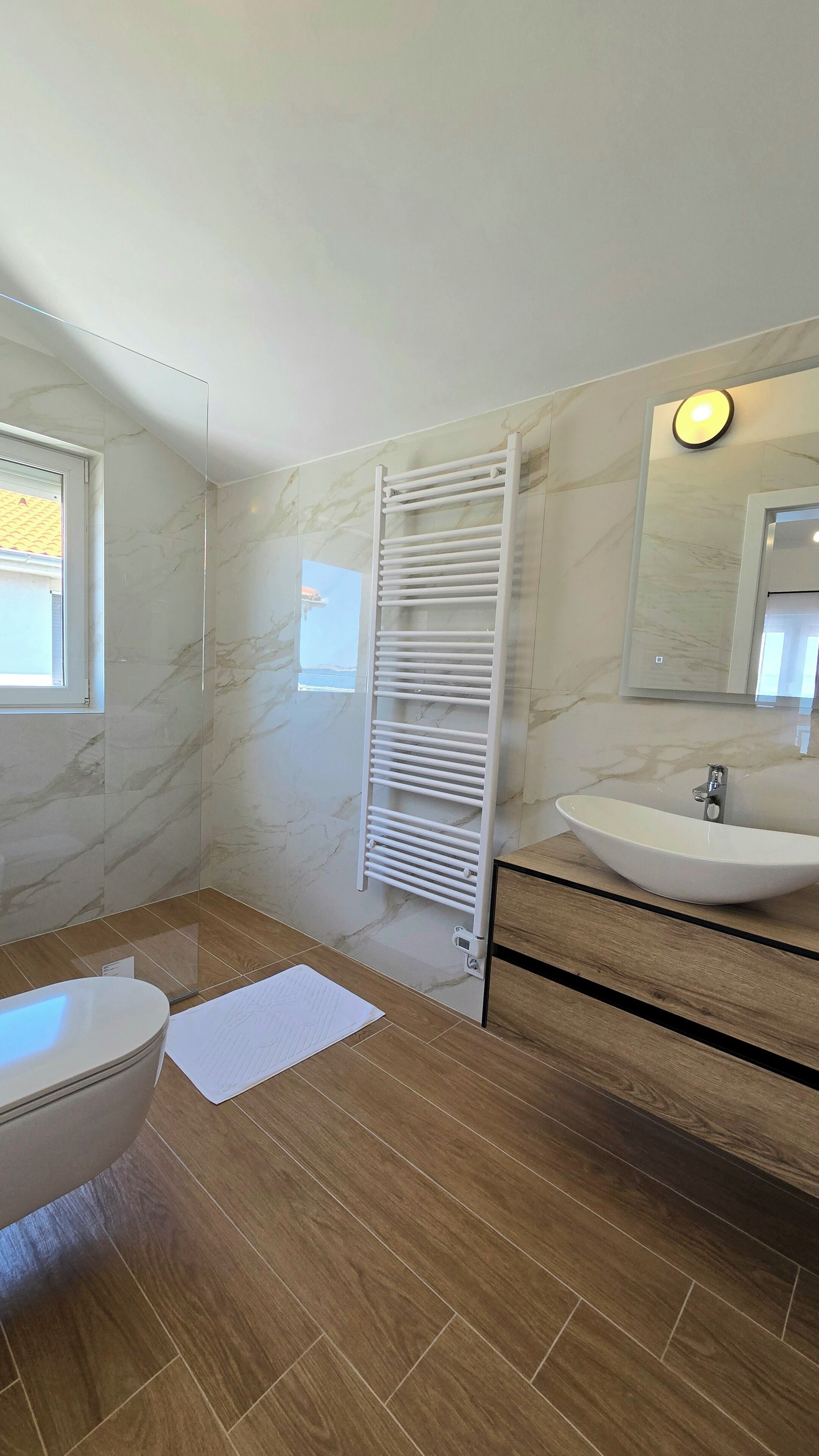 Un baño con pared de mármol, lavamanos blanco, suelo de madera y un perchero para toallas.