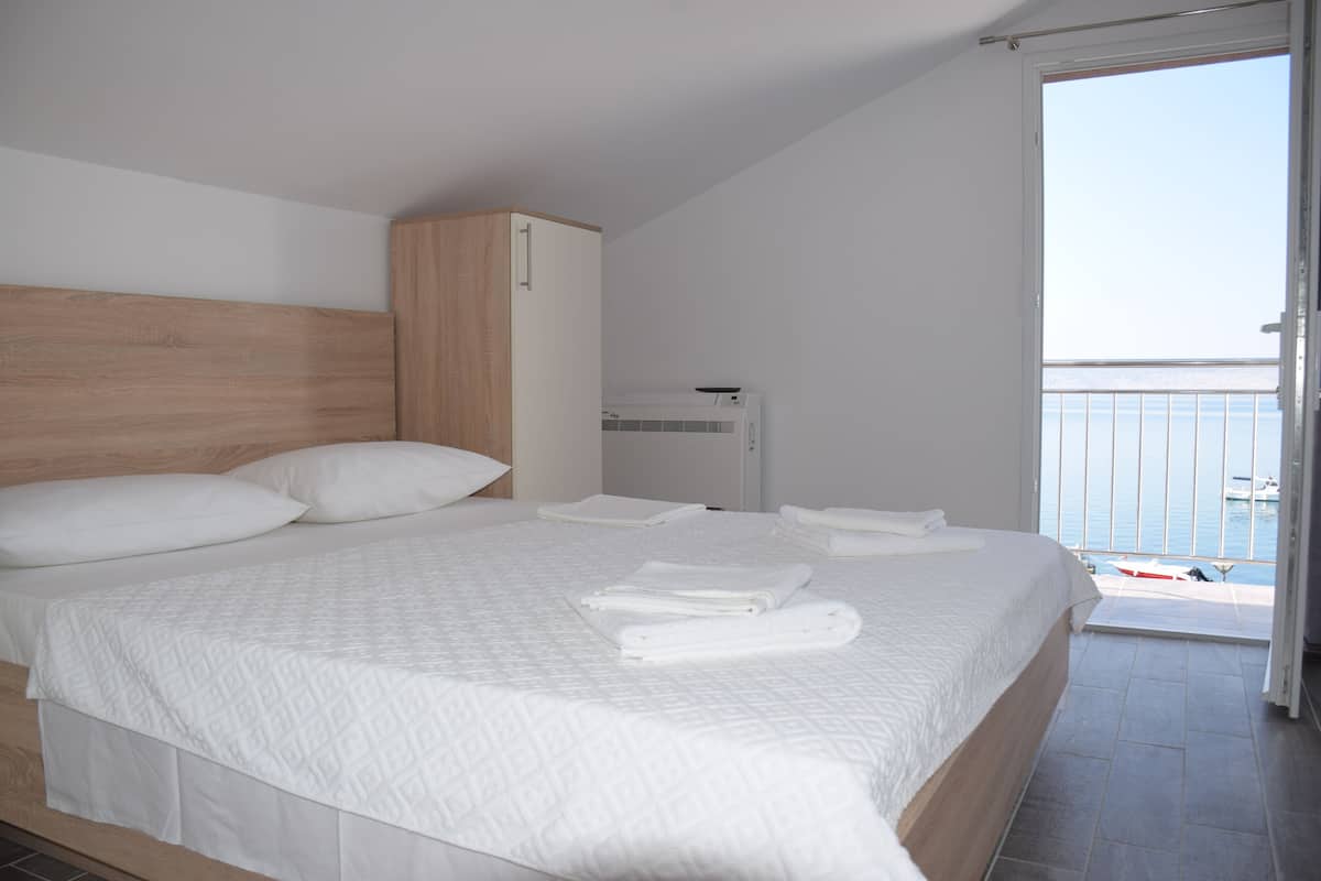 Une chambre avec un grand lit, une tête de lit en bois, une armoire blanche et un balcon avec vue sur la mer.