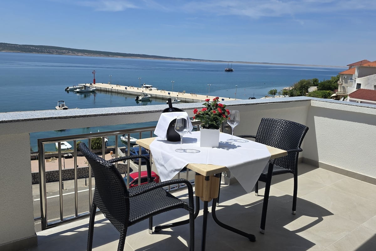 Un balcon avec une table dressée pour deux personnes, donnant sur une marina avec des bateaux et offrant une vue sur la côte.