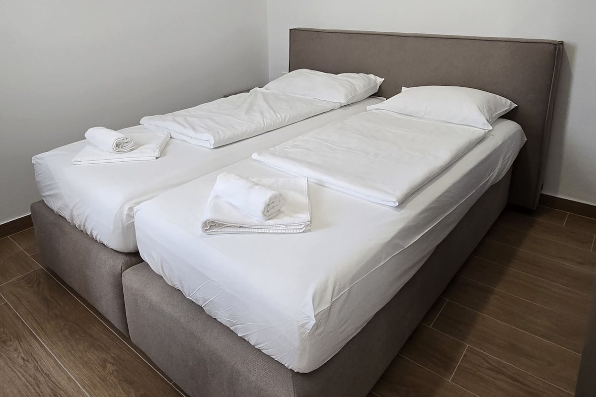 Un lit double avec du linge de lit blanc, une tête de lit grise et un plancher en bois.