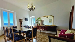 Spiseområde med et glasbord, træstole, en lysekrone, et ur, et fjernsyn og udsigt gennem vinduet.