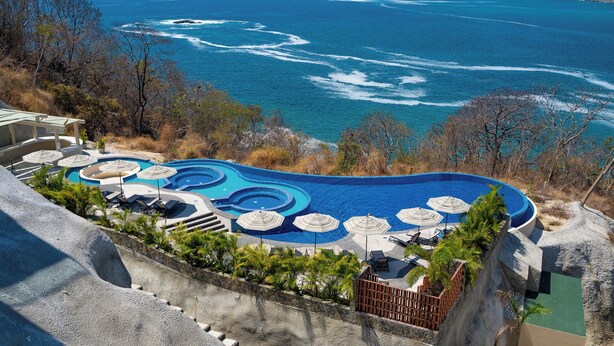 A swimming pool with a view of the ocean, surrounded by lounge chairs and umbrellas. - Stunning 2nd-Level | Views of Ocean, Beach & Pool (Huatulco)