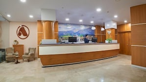 Zona de recepción de un hotel con un mostrador de madera, dos monitores de ordenador, un mural grande y un recepcionista detrás del mostrador.