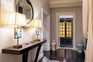 Un pasillo con una consola de madera oscura, dos lámparas, un espejo y una puerta que da a una habitación con suelo a cuadros.
