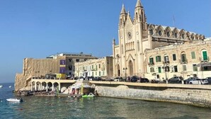 Ein Küstengebäude mit einem markanten Turm, einer Uferpromenade und Menschen am Strand.