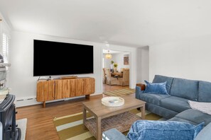A living room with a flat-screen TV, wooden cabinet, sofa, coffee table, and a dining area in the background.