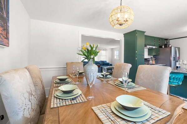 A dining area with a wooden table set for six, a vase with flowers, and a kitchen in the background.