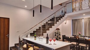 A modern kitchen with a central island, bar stools, a staircase, and a dining area with chairs and a table. - ELIVAAS Horizon Luxe 5BHK Villa (Udaipur)