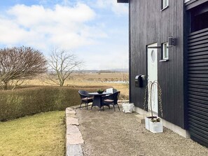 Een modern gebouw met een donkere gevel, een klein terras met een tafel en stoelen, en uitzicht op een landelijk landschap.