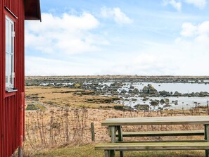 Een rood gebouw met een raam, een houten picknicktafel en uitzicht op een rotsachtige kustlijn met water.