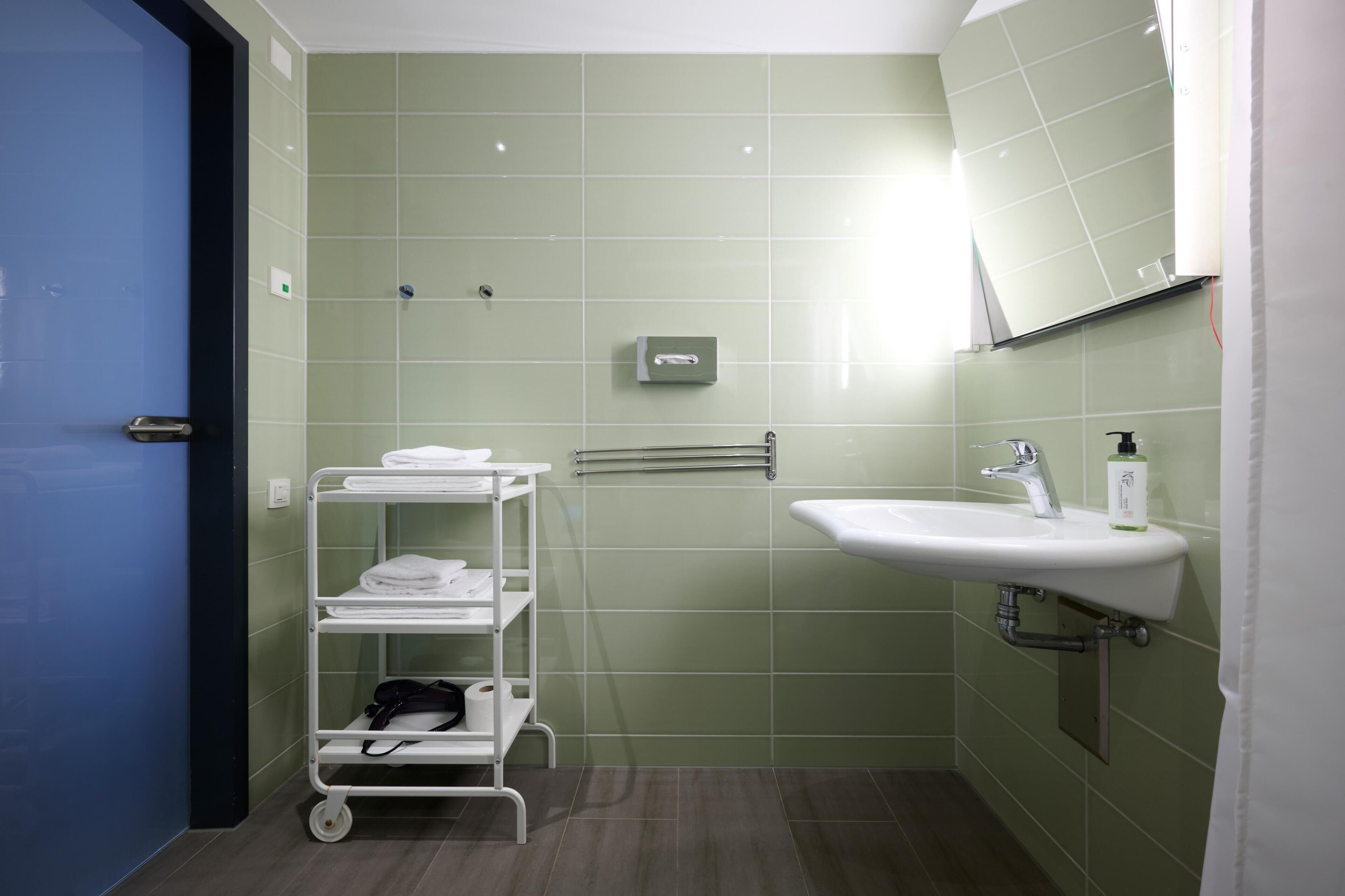 A bathroom with green tiles, a white sink, a mirror, a towel rack, and a cart with towels.