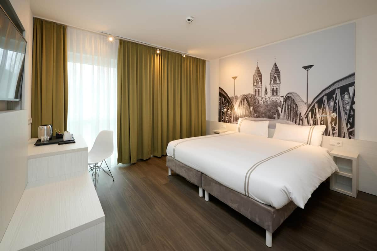 Une chambre d’hôtel comprenant un lit, un bureau, une chaise, une table de chevet et un mur orné d’une fresque représentant un paysage urbain.