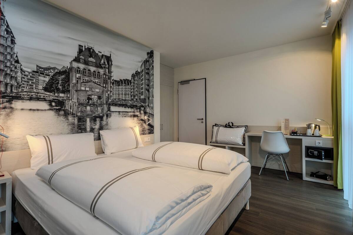 Ein Hotelzimmer mit Bett, Schreibtisch, Stuhl und einer großen Wandmalerei einer Stadtansicht.