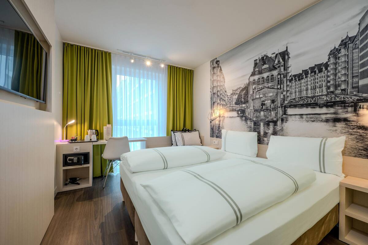 Ein Hotelzimmer mit einem Bett, einem Schreibtisch, einem Sessel, einem Fernseher und einer großen Stadtansicht als Wandgemälde.