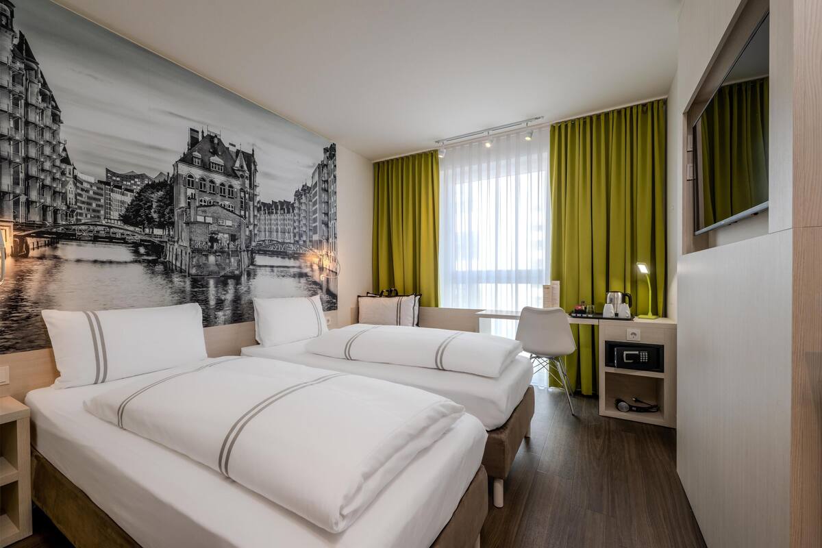 Ein Hotelzimmer mit zwei Betten, einem Schreibtisch, einem Sessel, einem Fernseher und einer großen Stadtansicht als Wandgemälde.