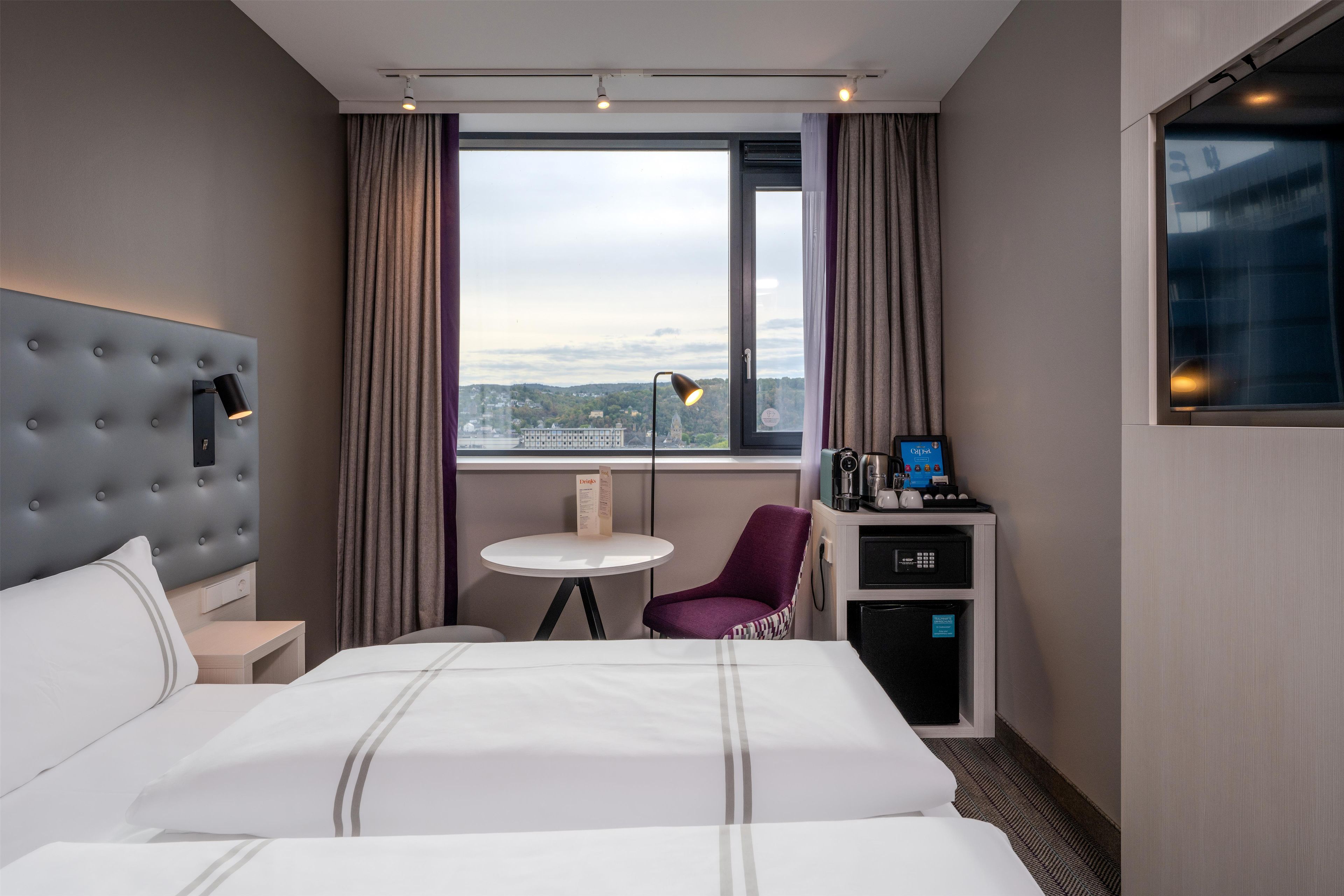 A hotel room with a large bed, a small table, a chair, a desk with a computer, and a window with a city view.