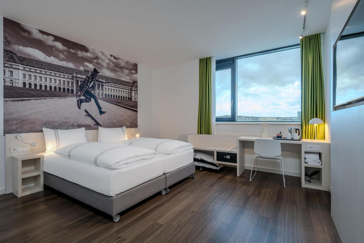 Habitación de hotel moderna con una cama grande, un escritorio, una silla, una lámpara de escritorio y vistas de la ciudad a través de un ventanal amplio.