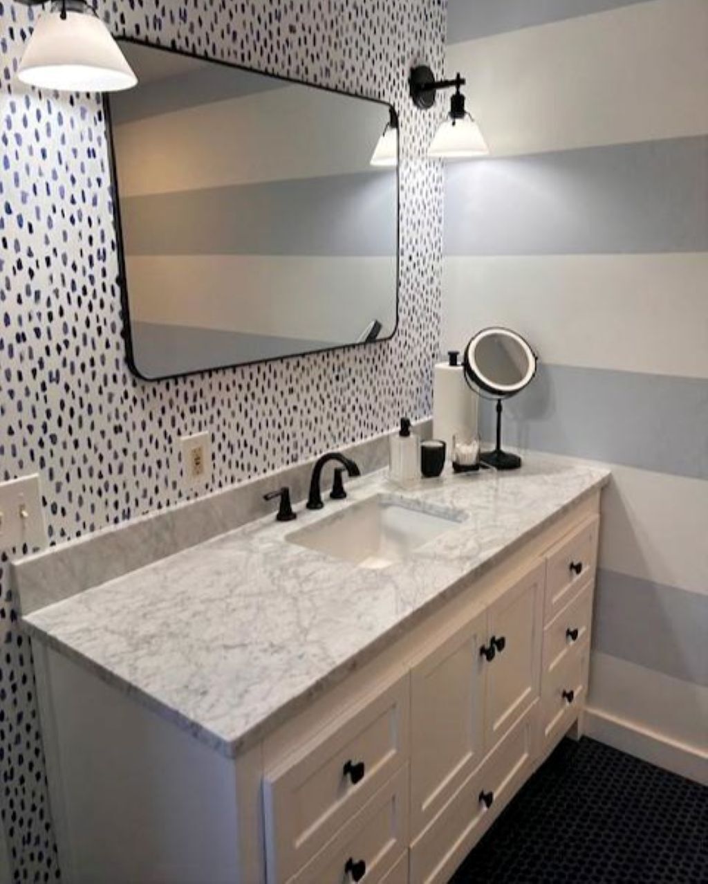 Une salle de bain avec un lavabo en marbre, deux robinets noirs, un grand miroir et un miroir rond posé sur le comptoir.