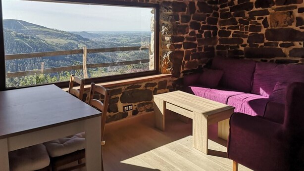Een rustieke kamer met een stenen muur, een houten plafond, een groot raam met uitzicht op het landschap, een houten tafel en een paarse bank.
