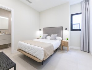 Quarto moderno com uma cama grande, mesas de cabeceira, um banco, uma janela e um banheiro visível por uma porta aberta.