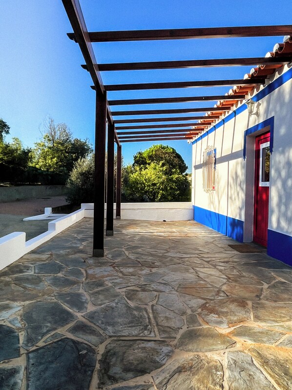 Une cour dallée de pierres, agrémentée d’une pergola, un bâtiment avec une porte rouge et un arbre en arrière-plan.
