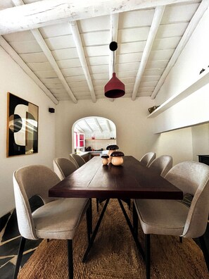 Uma sala de jantar com uma longa mesa de madeira, seis cadeiras, uma luminária pendente vermelha e vista para uma cozinha através de uma porta em arco.