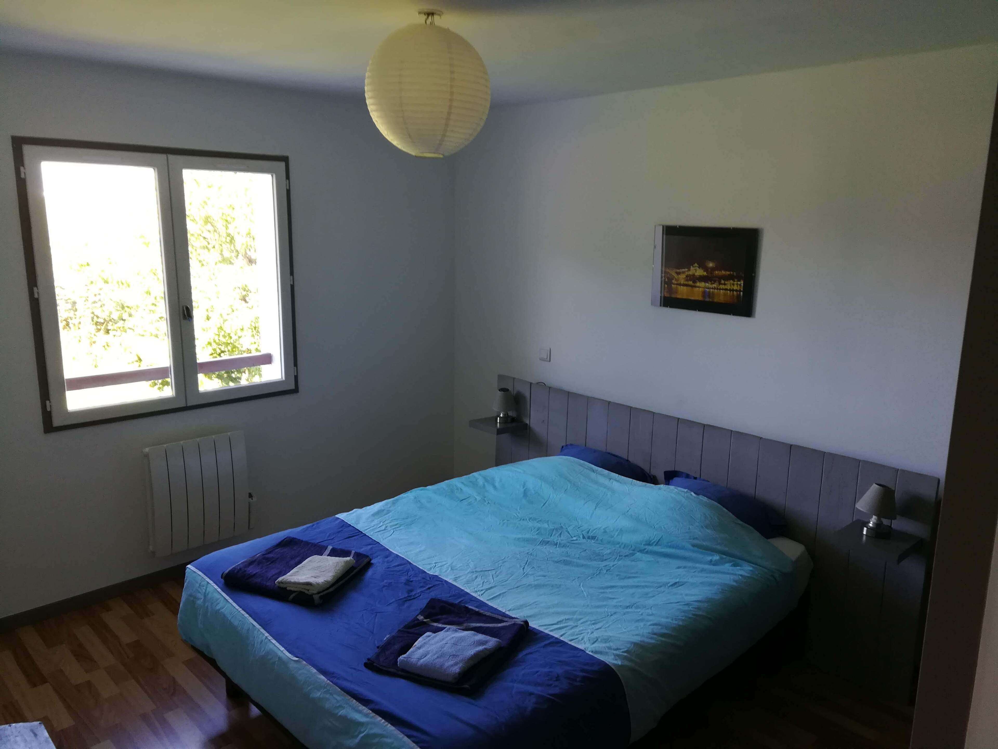 A bedroom with a bed, a window, a radiator, a wall-mounted lamp, and a framed picture.