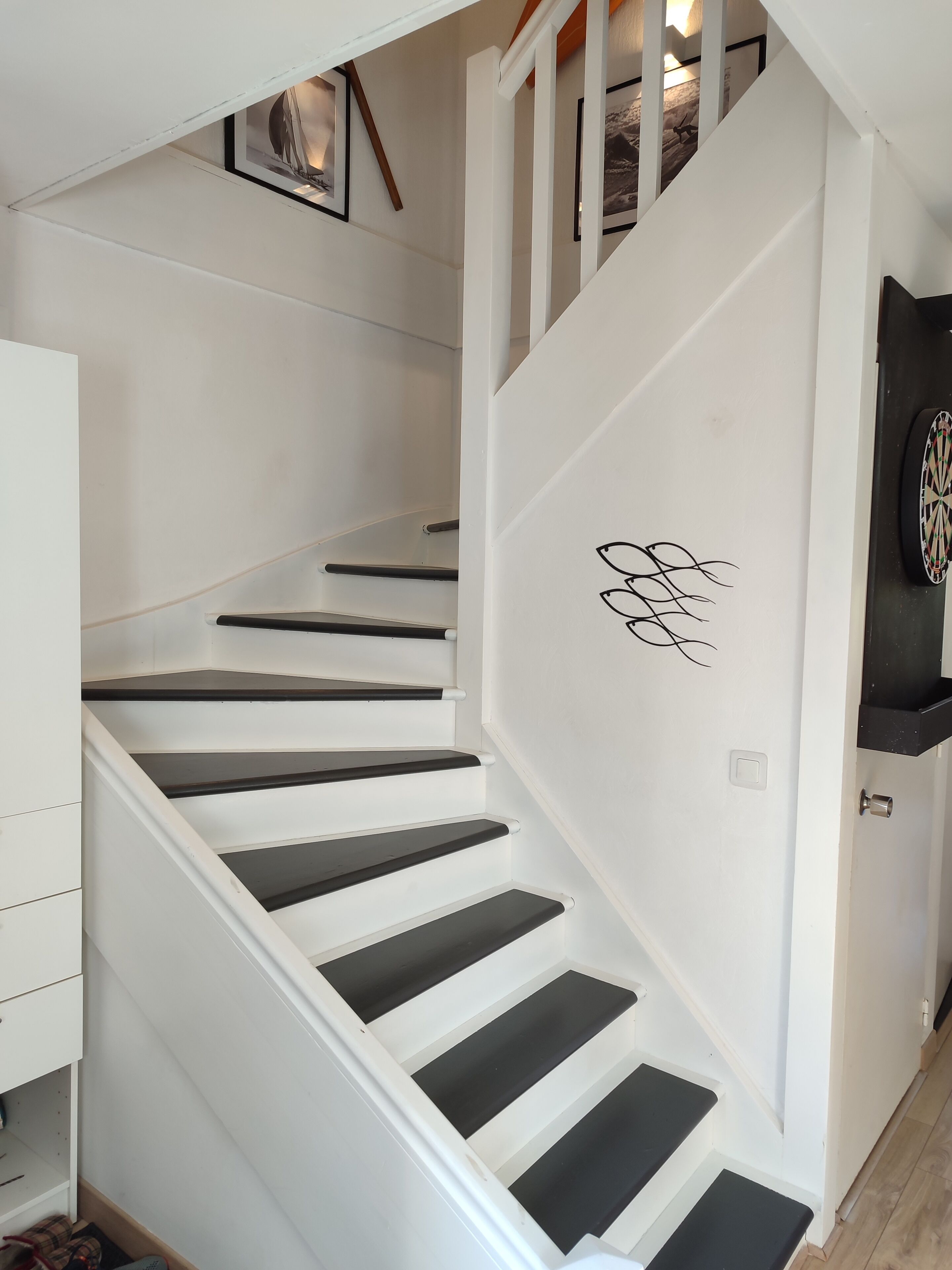 Un escalier blanc avec des marches noires, une porte ornée de motifs décoratifs et un jeu de fléchettes accroché au mur.
