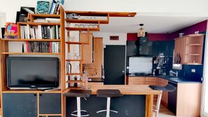 A compact living space with a kitchenette, a bar area with stools, a television, and a bookshelf filled with books and decorative items. - Cosy and quiet house between Landes and Pays Basque (Tarnos)