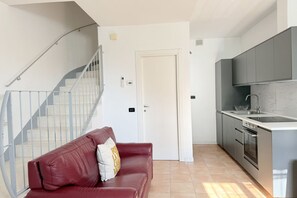 A modern kitchen with grey cabinets, a stainless steel sink, and an oven. There is a red leather sofa with a pillow in the living area. A white door is visible, leading to another room. - SE231 – Senigallia, renovated apartment with a/c (Senigallia)