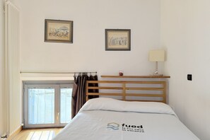 A hotel room with a bed, a window with curtains, a framed picture on the wall, and a small table with a lamp.