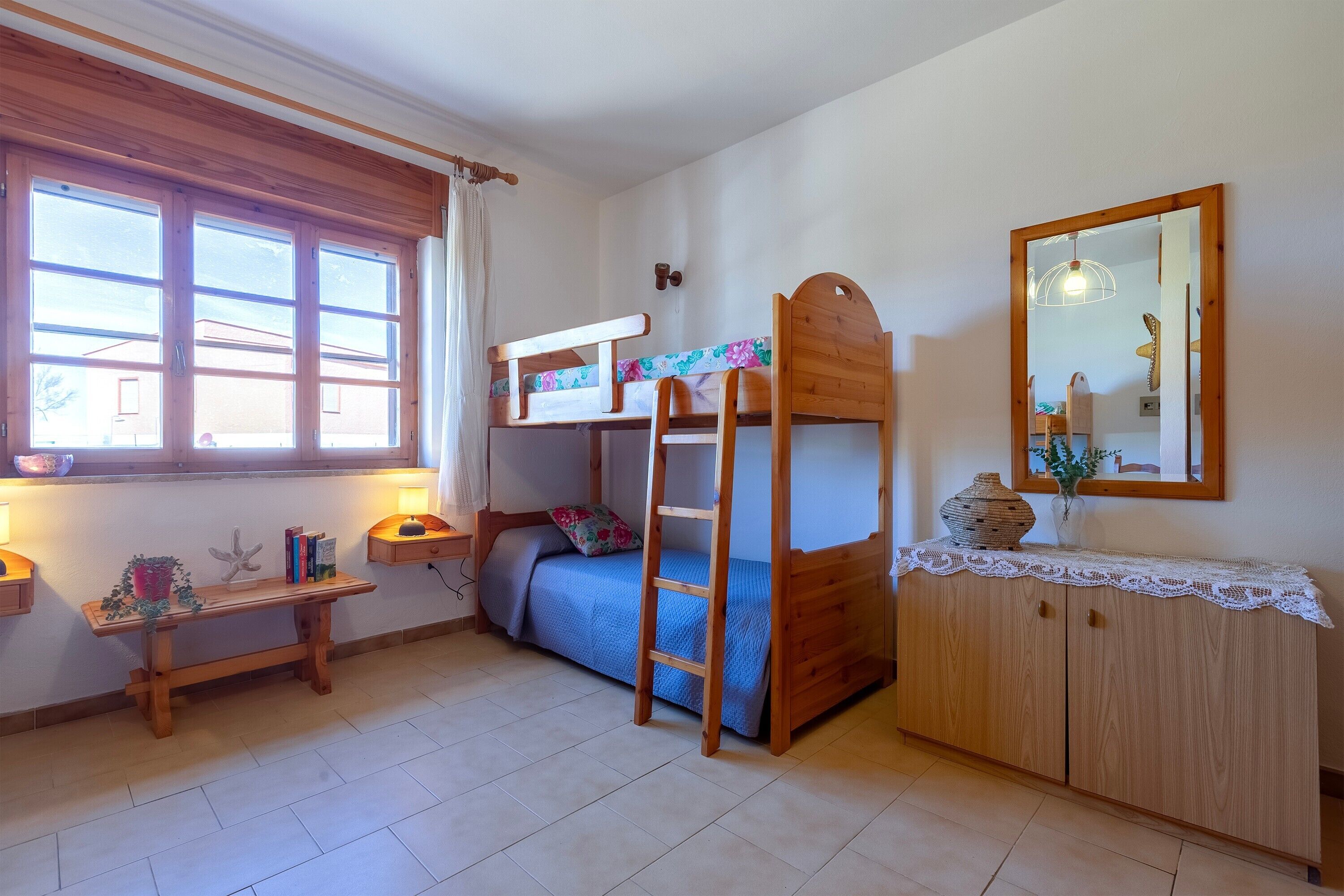 A bedroom with a bunk bed, a bedside table, a lamp, a mirror, and a window with curtains.