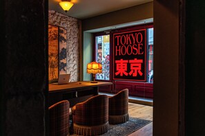 Et værelse med et træskrivebord, en lampe og ternede stole. En neonskilte viser 'TOKYO HOOSE' på engelsk og japansk.