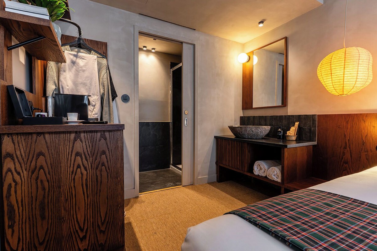 A hotel room with a wooden dresser, a large mirror, a bowl sink, a bed with a plaid blanket, and a hallway leading to a bathroom.