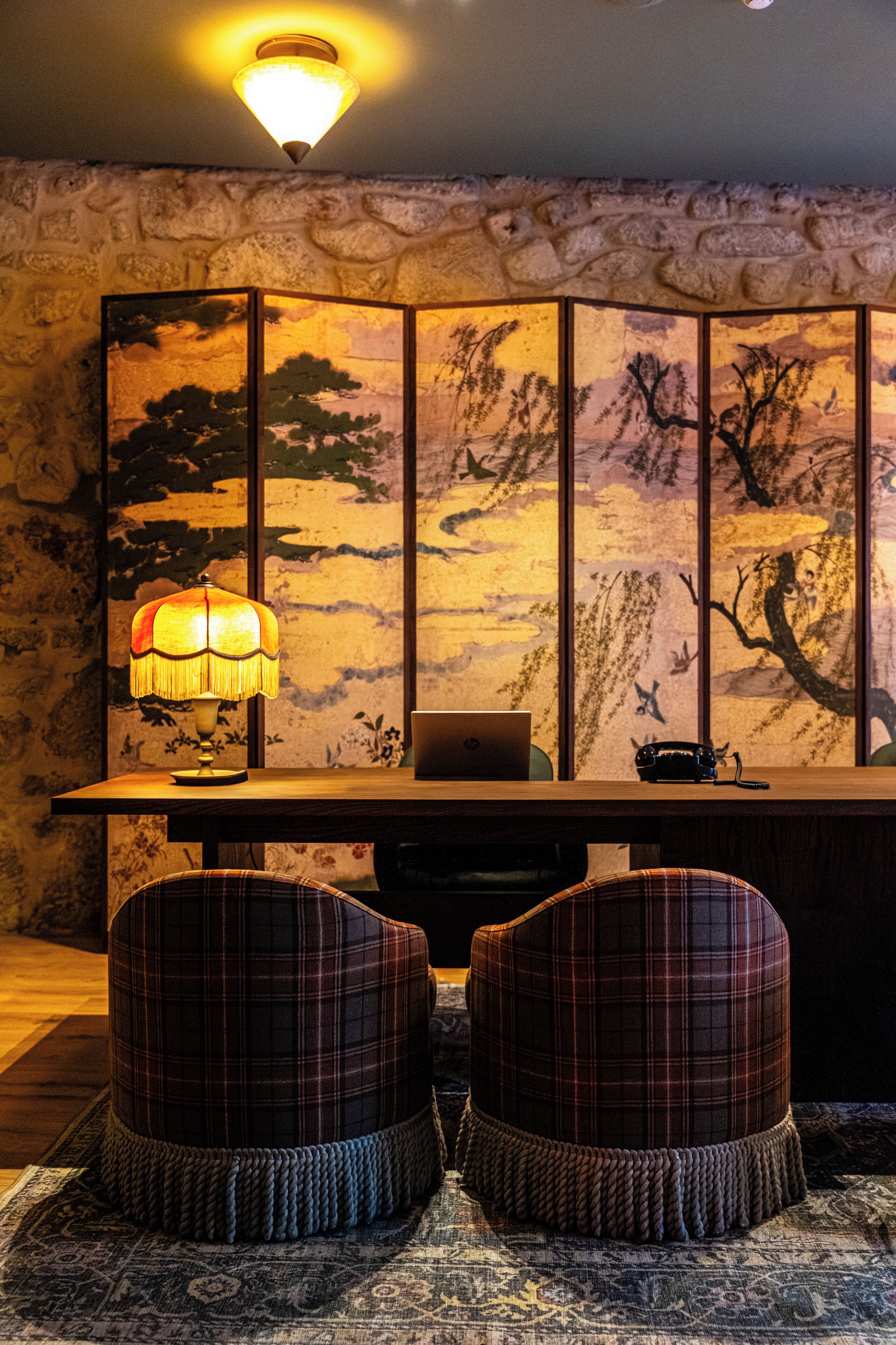 A room with a wooden desk, a laptop, a lamp, and two plaid chairs.
