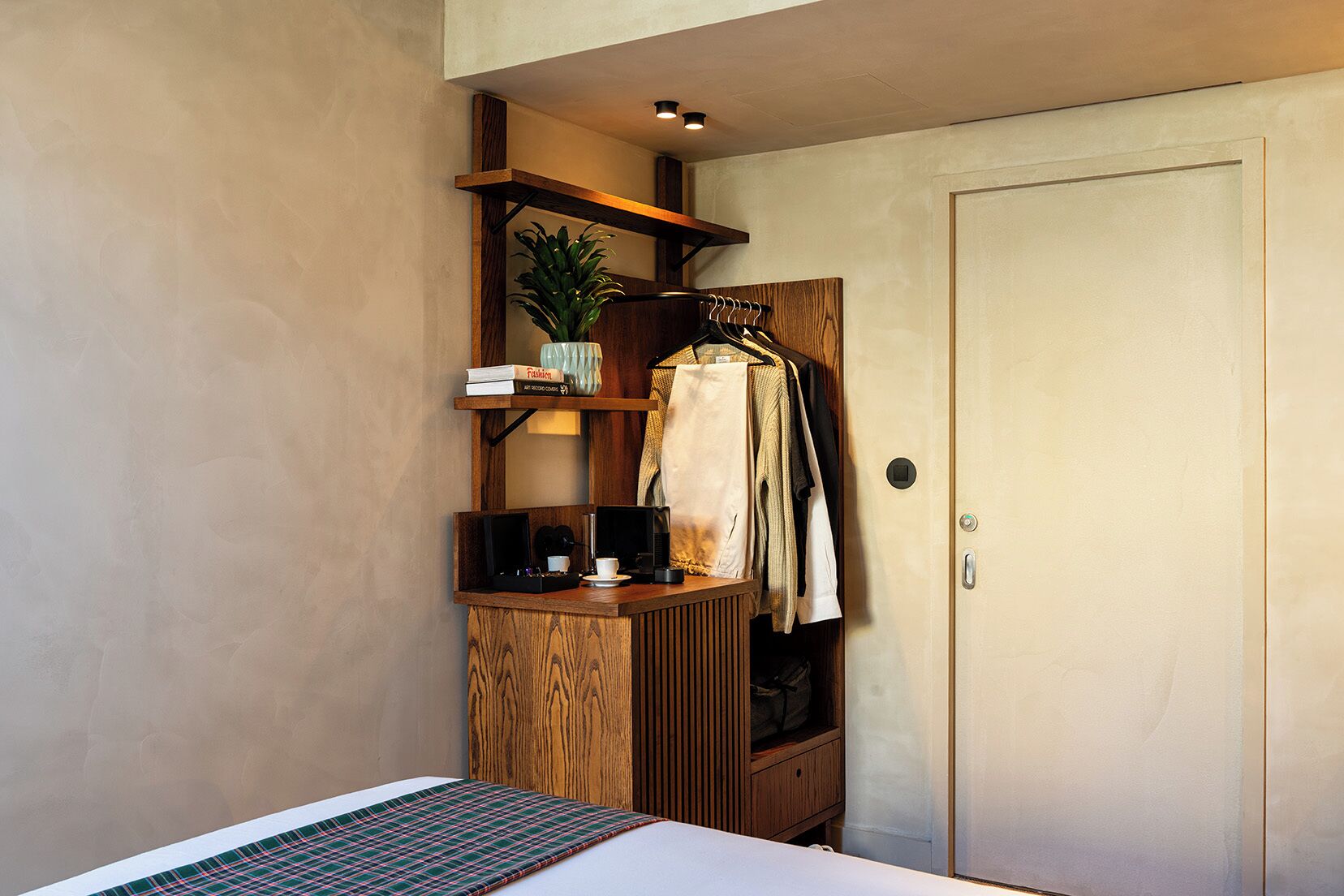 A hotel room with a bed, a wooden side table with a coffee machine, a hanging coat rack, and a door with a lock.