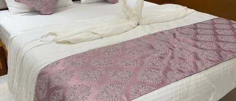A neatly made bed with a white and pink patterned bedspread, a heart-shaped towel decoration, and a headboard with multiple pillows.