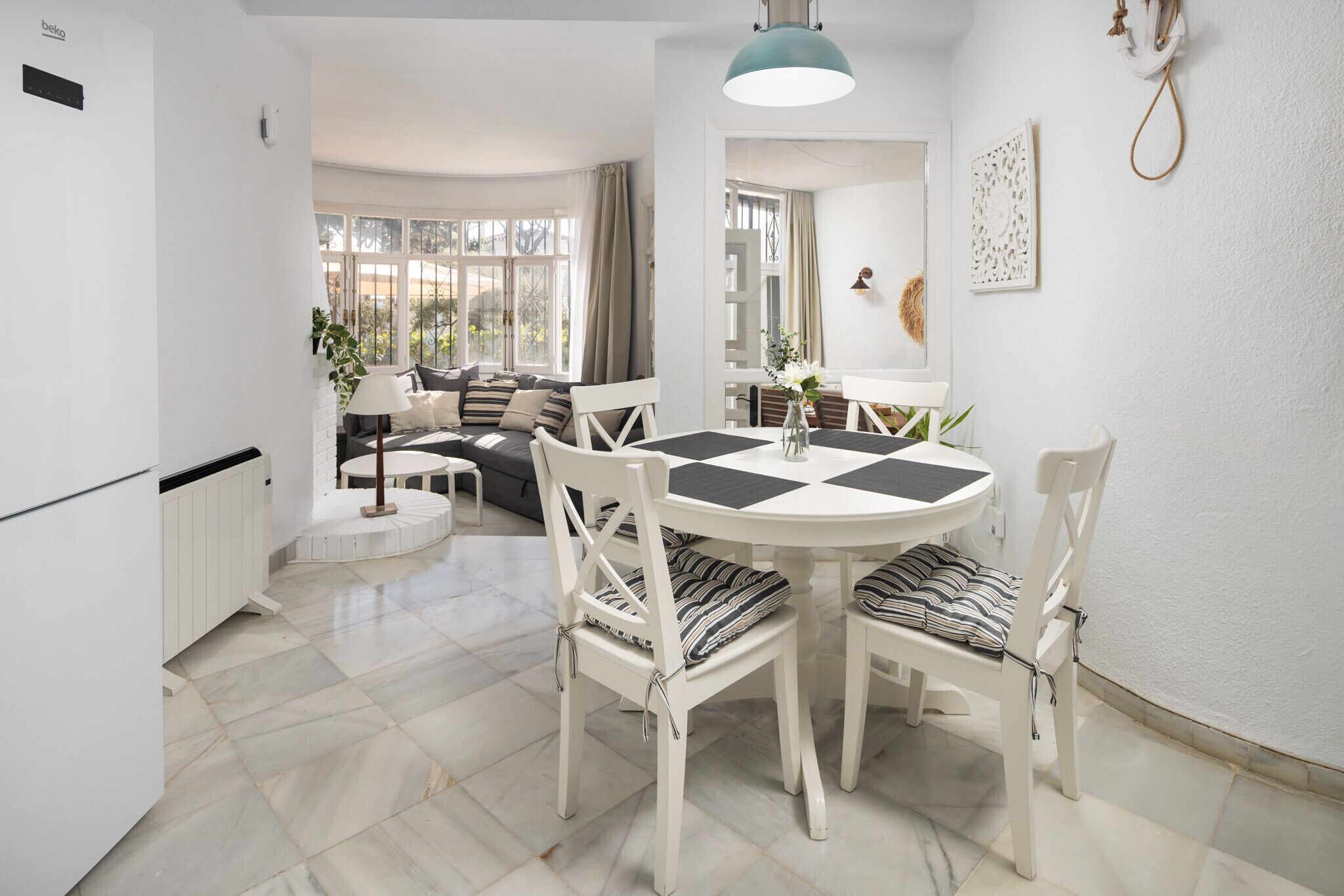 A dining area with a white table and chairs, a pendant light, and a view of the outdoors through large windows.