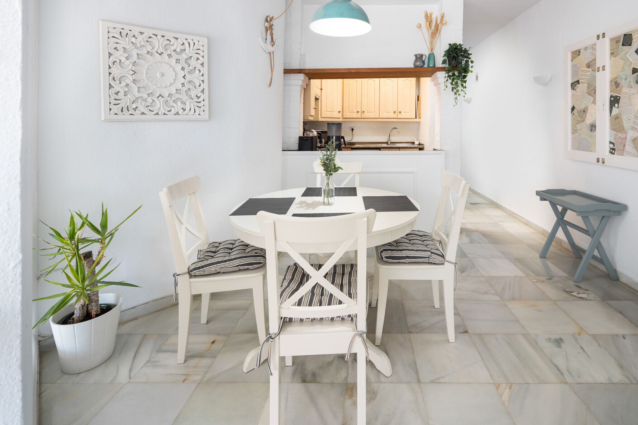 A dining area with a round table, white chairs, a potted plant, and a kitchen in the background.