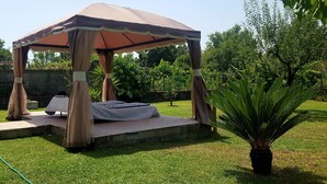 Un mirador con dosel, una mesa y sillas, rodeado de césped y árboles.