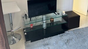A modern living room with a flat-screen TV on a glass shelf, a white rose painting on the wall, a glass coffee table, a black ottoman, and a white chair. - Comfortable apartment with balcony and huge pool (Sucina)